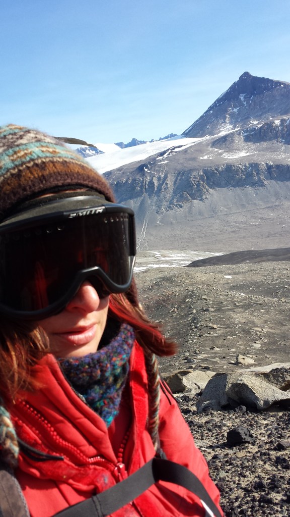 A woman with medium length pick hair wearing snow goggles, a heavy read coat, a hat, and a scarf in front of barren mountains and a glacier