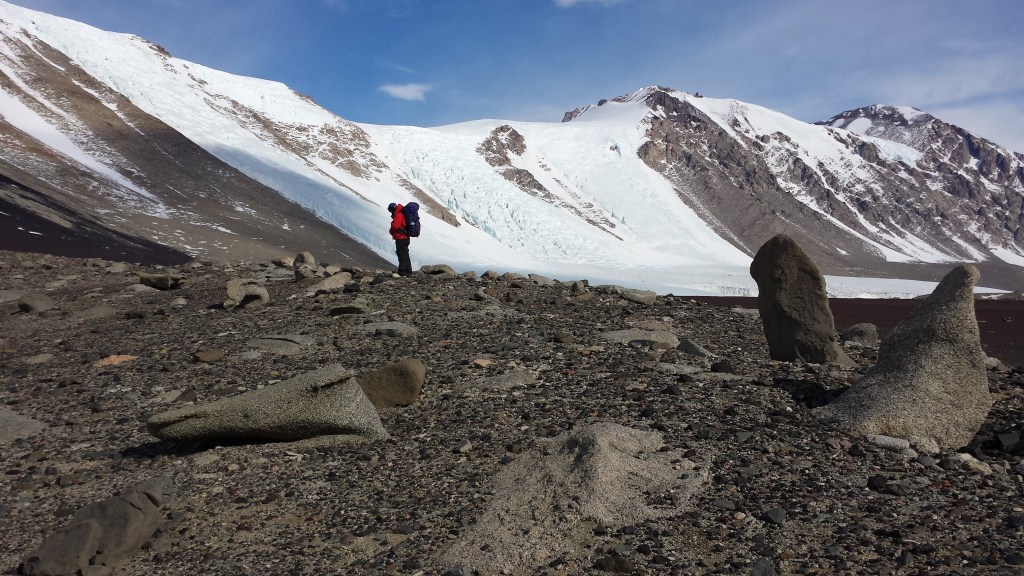 A landscape with glaciers and barren mountains, and rocks in the foreground. A small person with a big backpack stands in the middle distance